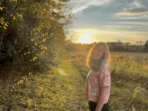 Rebecca Doris stands in the meadows with a sunset in the background, facing the camera and smiling laughing.