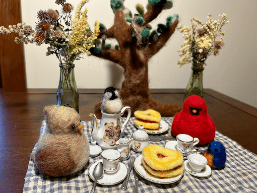 Assorted needle felting critters have a tea party on a picnic blanket. From left to right, a female Northern Cardinal borb, a mini black-capped chickadee sits in a teapot, a male Northern Cardinal, and a mini Eastern bluebird. Needle-felted heart-shaped raspberry jam cookes lie on the picnic plates.