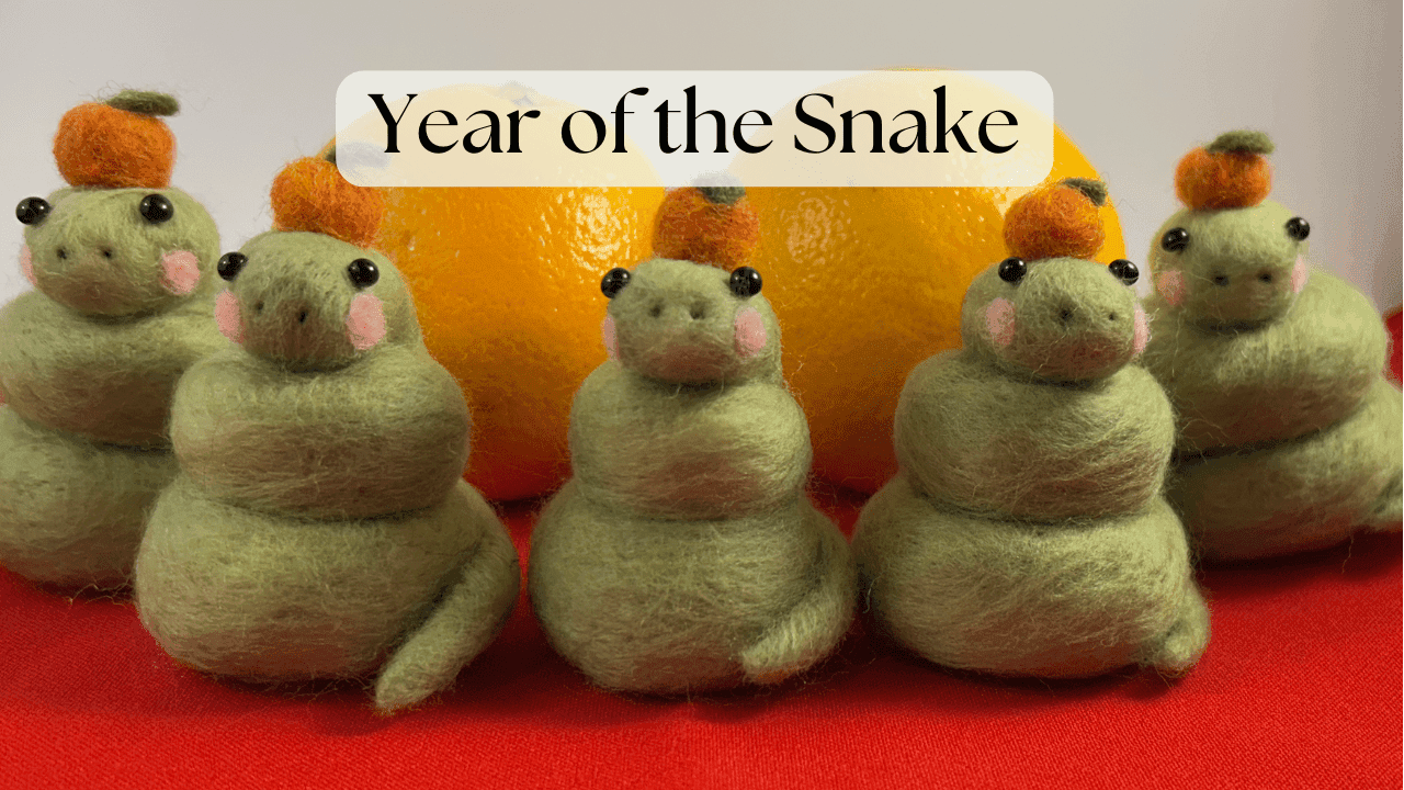 Five needle-felted snakes sit on a red cloth in front of two orange. The snakes are light green with pink cheeks and wearing mandarin oranges on their heads.