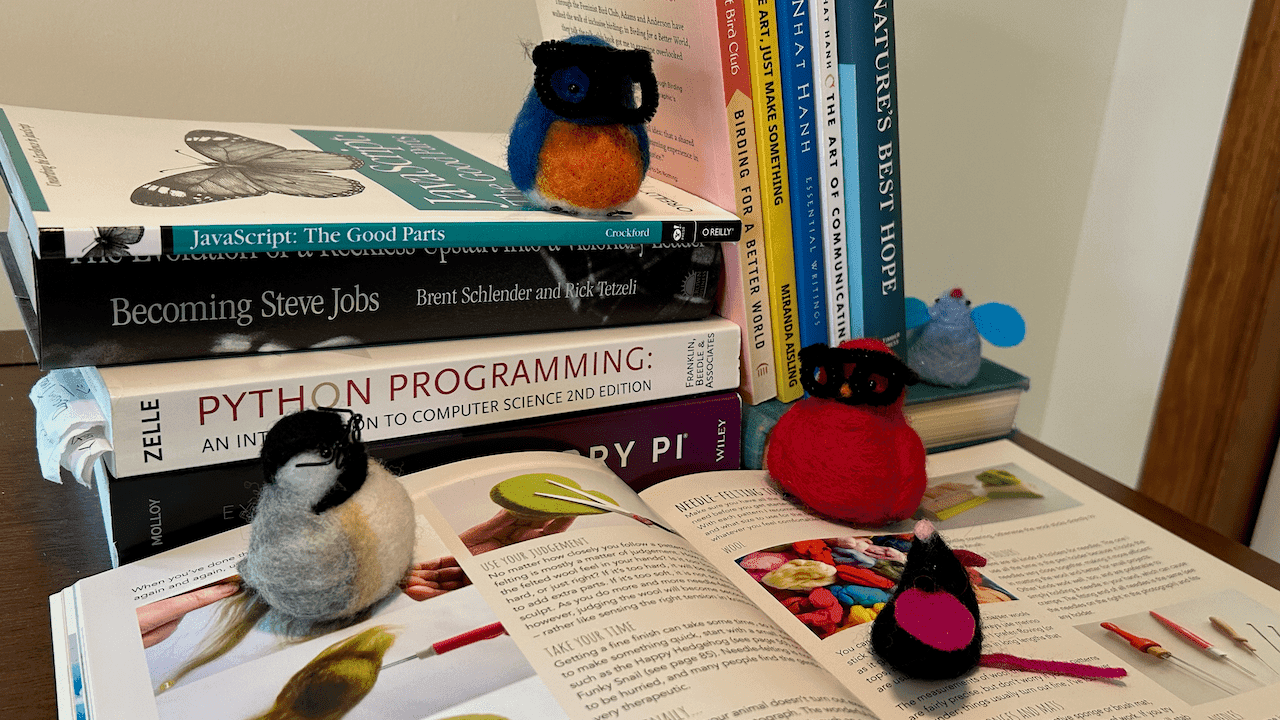 Assorted needle-felted critters study the book _Little Needle Felt Animals_ by Gretel Parker. Clockwise from left, there's a bespectacled black-capped chickadee borb, a male Eastern bluebird borb wearing pipe cleaner glasses sitting on a stack of books, a male Northern cardinal borb also wearing pipe cleaner glasses, a blue little mouse sitting next to some leaning books, and a black little mouse. The stack of books in the background is: _Exploring Raspberry Pi_ by Derek Molloy, _Python Programming: An Introduction to Computer Science 2nd Edition_by Zelle, _Becoming Steve Jobs_ by Brent Schlender and Rick Tetzeli, and  _JavaScript: The Good Parts_ by Crockwell. The leaning books from left to right are _Birding for a Better World_ by Feminist Bird Club, _Don't Make Art, Just Make Something_ by Miranda Aisling, _Essential Writings_ by Thich Nhat Hanh, _The Art of Communication_ by Thich Nhat Hanh, and _Nature's Best Hope_ by Douglass Tallamy.