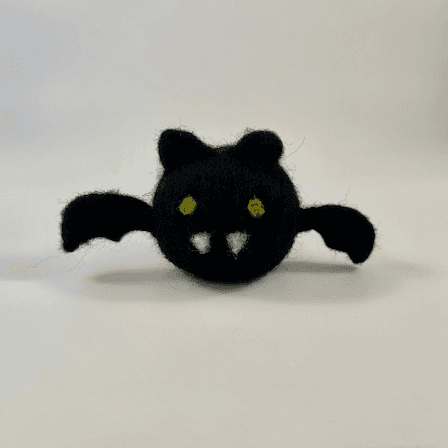 A needle-felted black bat with a round orb-like body faces forward. It has green eyes, two white pointed teeth, round ears, and small wings.