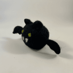 A needle-felted black bat with a round orb-like body faces 3/4 to the left. It has green eyes, two white pointed teeth, round ears, and small wings.