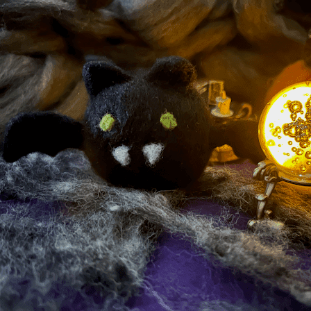 A needle-felted black bat with a round orb-like body faces forward. It has green eyes, two white pointed teeth, round ears, and small wings. It sits next to a glowing crystal ball in a spooky scene with a purple and cob web floor and dingy background.