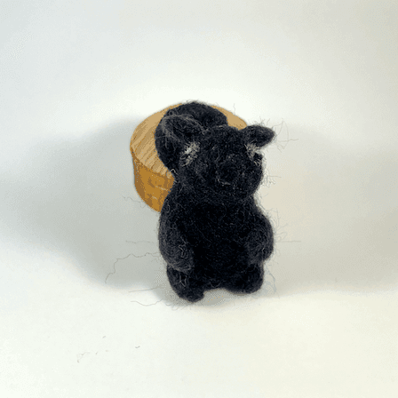 A needle-felted black squirrel faces directly towards the camera. Its tail rests on a circle of cut wood behind it.