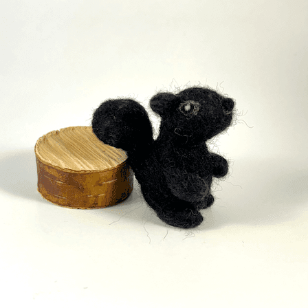 A needle-felted black squirrel faces directly towards the right. Its tail rests on a circle of cut wood behind it.