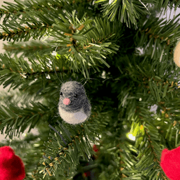 A close up of a needle-felted dark-eyed junco ornament, hanging on a tree, facing 1/4 to the left.