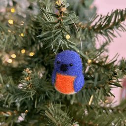 A close up of a needle-felted Eastern bluebird hanging from a Christmas tree, facing slightly towards the left.