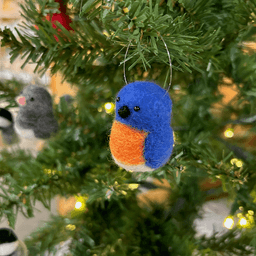 A close up of a needle-felted Eastern bluebird hanging from a Christmas tree, facing 3/4 towards the left.