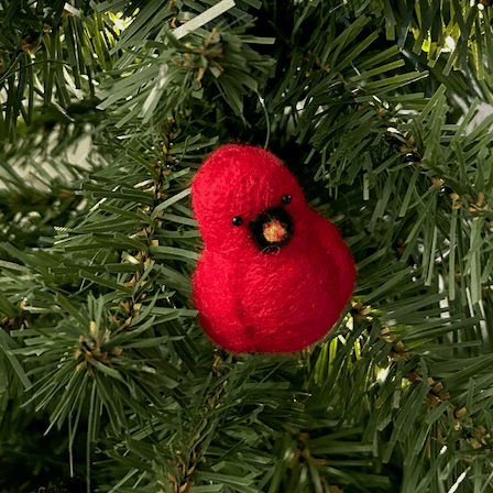 A close up of a needle-felted Northern cardinal ornament hanging on a tree, facing slightly to the right.