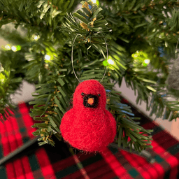 A close up of a needle-felted Northern cardinal ornament hanging on a tree, facing directly towards the camera.