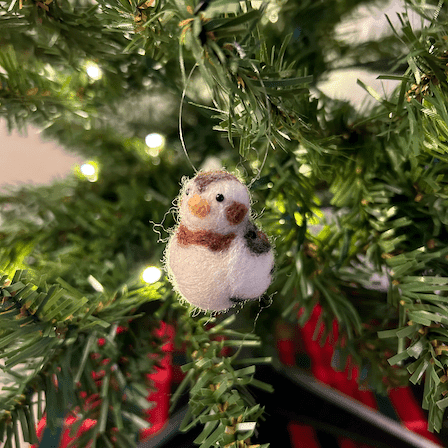 A close up of a needle-felted snow bunting ornament hanging on a tree, facing 1/4 to the left.