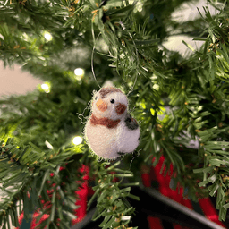 A close up of a needle-felted snow bunting ornament hanging on a tree, facing 1/4 to the left.