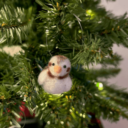 A close up of a needle-felted snow bunting ornament hanging on a tree, facing directly towards the camera.