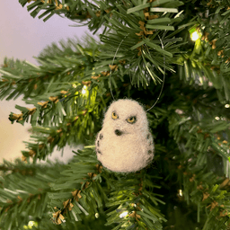 A close up of a needle-felted snowy owl ornament hanging on a tree, facing slightly to the left. Its wings have bands.