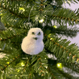 A close up of a needle-felted snowy owl ornament hanging on a tree, facing slightly to the right. The owl is completely white.