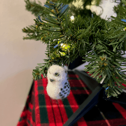 A close up of a needle-felted snowy owl ornament hanging on a tree, facing 1/4 to the left. Its wings and the back of its head have bands.