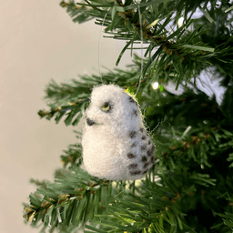 A close up of a needle-felted snowy owl ornament hanging on a tree, facing 1/4 to the left. Its wings have bands.