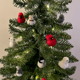 Close up of a small artificial Christmas tree with white lights. The tree's ornaments are all borbs: two snowy owls, one black-capped chickadee, two Northern cardinals, one snow bunting, two dark-eyed juncos. The snow bunting is in the top left to the right of the cardinal.