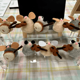 Assorted needle-felted mice and possums playing fiddles and banjos sit on a clear two-level shelf on a crafting display table.