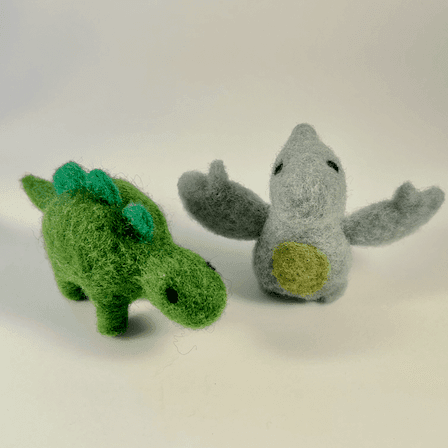 Two needle felted dinosaurs, a green stegasaurus and a grey blue pterodactyl, stand side by side. The stegasaurus on the left faces 1/4 towards the right. The pterodactyl on the left faces directly towards the camera.