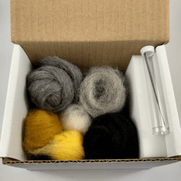 Inside a small open cardboard box, small skeins of rolled up Charcoal fiber, Shadow fiber, Rust fiber, Sunshine fiber, Snowball fiber, and Black fiber. On top a felting needle inside a plastic tube. On the right against the box wall, a foam block felting pad.