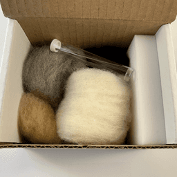 Inside a small open cardboard box, small skeins of rolled up Snowball fiber, Buff fiber, Charcoal fiber, and Black fiber. On top a felting needle inside a plastic tube. On the right against the box wall, a foam block felting pad.