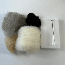 Laid out are small skeins of rolled up Snowball fiber, Buff fiber, Charcoal fiber, and Black fiber. To the right, a foam block felting pad on top of which is a tube containing a felting needle.