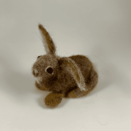 A needle-felted Eastern cottontail rabbit faces 3/4 towards the left. Its ears are splayed widely. Its cheeks are round, its nose small, and its large dark eyes bulbous.