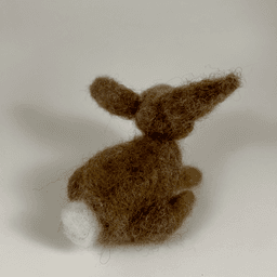 A needle-felted Eastern cottontail rabbit faces away from the camera, 1/4 towards the right, showing its brown back and cotton puff tail.