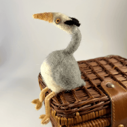 A needle-felted great blue heron borb with pipe cleaner legs, facing towards the left, sits on a small wicker case.