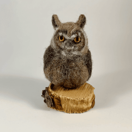 A needle-felted great horned owl sits on a small circle of cut wood, facing directly towards the camera.