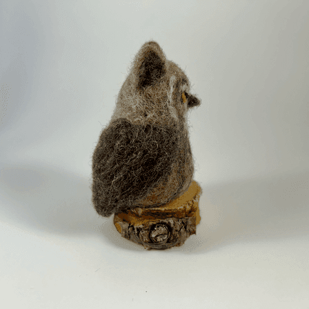 A needle-felted great horned owl sits on a small circle of cut wood, facing towards the right.