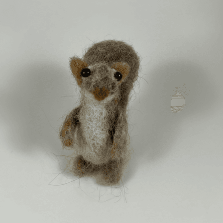 A mostly brown-colored Eastern grey squirrel, sculpted in needle-felt, faces towards the camera. Its nose is reddish brown, its belly, neck and under-nose area are cream colored, its ears and paws are a lighter reddish brown. The rest of its body is a greyish brown. Its eyes are black beads.