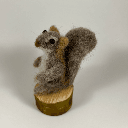 A mostly brown-colored Eastern grey squirrel, sculpted in needle-felt, standing on a round section of branch faces towards the left, displaying its rounded bushy grey-brown tail.