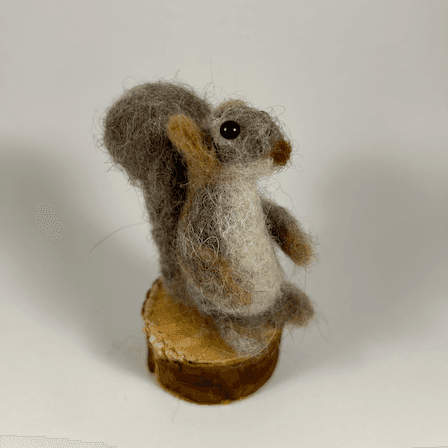 A mostly brown-colored Eastern grey squirrel, sculpted in needle-felt, standing on a round section of branch faces 1/4 towards the right, showing its profile, round snout and slightly curved cream colored belly.