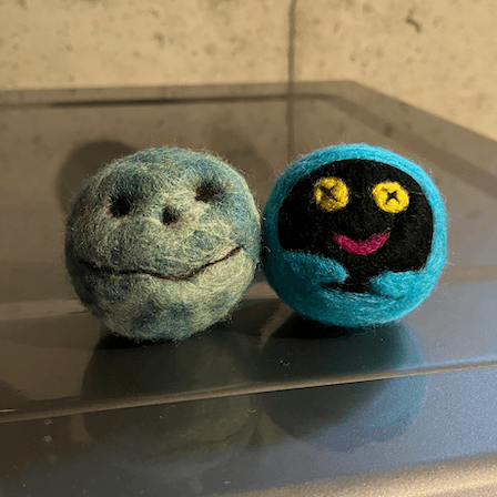 Sitting on top of a basement dryer, two needle felted dryer balls that resemble Chthonic Companion characters from the video game Hades. On the left is Bouldy, a smiling grey green and blue boulder. On the right is Shadey, a ghost plush with yellow button eyes, a pink smile, a black face, and a blue body and arms.