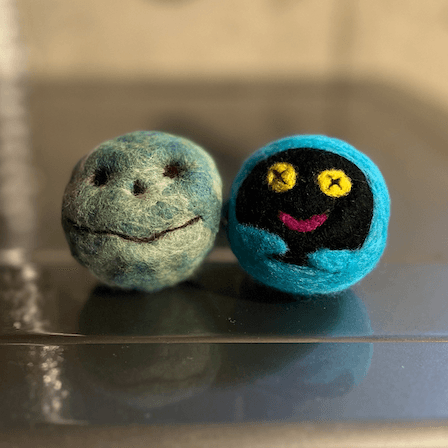 Sitting on top of a basement dryer, two needle felted dryer balls that resemble Chthonic Companion characters from the video game Hades. On the left is Bouldy, a smiling grey green and blue boulder. On the right is Shadey, a ghost plush with yellow button eyes, a pink smile, a black face, and a blue body and arms. A dryer vent tube reflects in the top of the dryer.