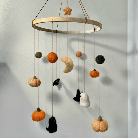 Full view of a Halloween baby mobile strung from an embroidery hoop. The mobile items include an orange star and yellow crescent moon, green and orange balls, pumpkins, a ghost, a witch hat, a black cat, and a bat.