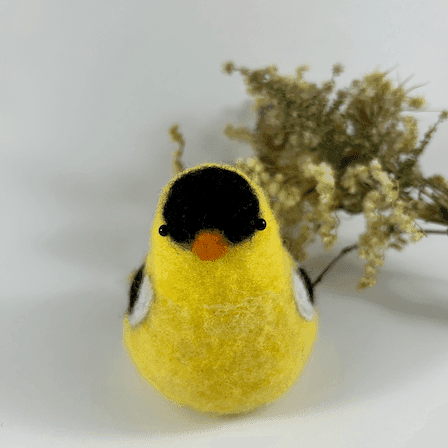 A needle-felted American goldfinch in breeding plumage, pear-shaped, with black bead eyes directly faces the camera. A spray of dried American aster and goldenrod lies in the background.