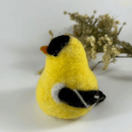 A needle-felted American goldfinch in breeding plumage, pear-shaped, with black bead eyes directly faces towards the left. A spray of dried American aster and goldenrod lies in the background.