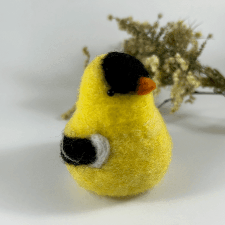 A needle-felted American goldfinch in breeding plumage, pear-shaped, with black bead eyes directly faces 1/4 towards the right. A spray of dried American aster and goldenrod lies in the background.