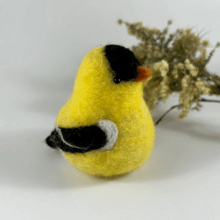 A needle-felted American goldfinch in breeding plumage, pear-shaped, with black bead eyes faces towards the right. A spray of dried American aster and goldenrod lies in the background.