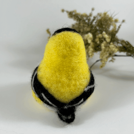 A needle-felted American goldfinch in breeding plumage, pear-shaped, with black bead eyes directly faces directly away from the camera. A spray of dried American aster and goldenrod lies in the background.