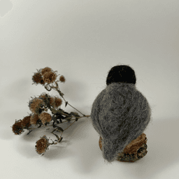 A needle-felted American Robin sits on a half inch section of a branch faces away from the camera. A dried spray of New England aster lies behind it. Its head is black, its back, wings and tails are grey.
