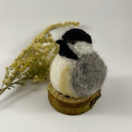 A needle-felted black-capped chickadee sits on a half inch section of a branch, facing 3/4 towards the left. A dried spray of goldenrod lays in the background. Its belly is white with buff sides, its face white, its goatee black, its beak black, its cap black, its wings grey.