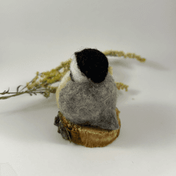 A small needle-felted black-capped chickadee sits on a half inch section of a branch, facing away from the camera 1/4 towards the left. A dried spray of goldenrod lays in the background. Its tail is grey, its back is grey, its wings are grey, its face white, its cap black.