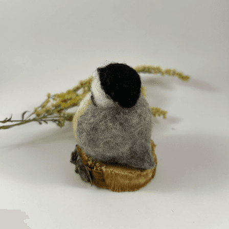 A small needle-felted black-capped chickadee sits on a half inch section of a branch, facing away from the camera 1/4 towards the left. A dried spray of goldenrod lays in the background. Its tail is grey, its back is grey, its wings are grey, its face white, its cap black.