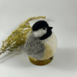 A needle-felted black-capped chickadee sits on a half inch section of a branch, facing 3/4 towards the right. A dried spray of goldenrod lays in the background. Its belly is white with buff sides, its face white, its goatee black, its beak black, its cap black, its wings grey.