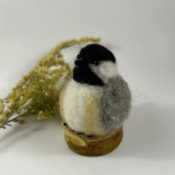 A needle-felted black-capped chickadee sits on a half inch section of a branch, facing 1/4 towards the left. A dried spray of goldenrod lays in the background. Its belly is white with buff sides, its face white, its goatee black, its beak black, its cap black, its wings grey.