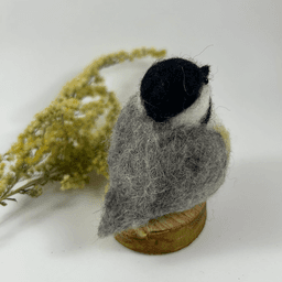 A needle-felted black-capped chickadee sits on a half inch section of a branch, facing away from the camera 1/4 towards the right. A dried spray of goldenrod lays in the background. Its tail is grey, its back is grey, its wings are grey, its face white, its cap black, its peeking breast buff and white.